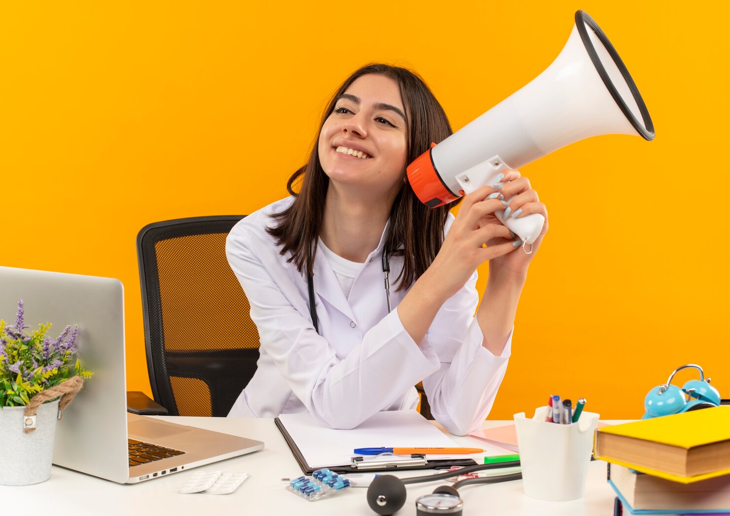 joven doctora bata blanca estetoscopio sosteniendo megafono sonriendo cara feliz sentado mesa computadora portatil documentos sobre pared naranja 141793 52496